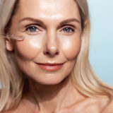 Close up of middle aged smiling woman, applies C-Serum with dropper, collagen anti-aging oil for skin lifting and glowing tone, blue background.
